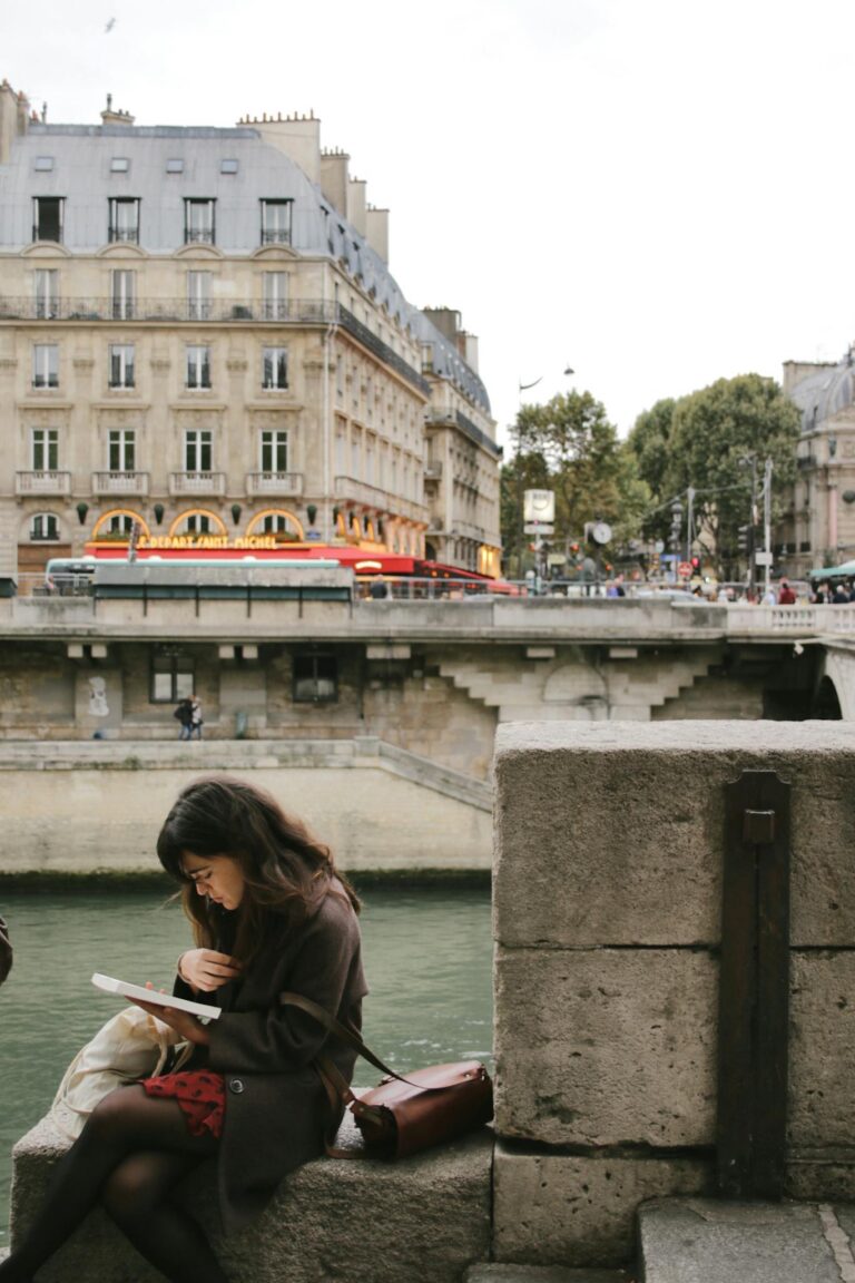 A woman reading a book by the Seine River with Parisian architecture in the background.